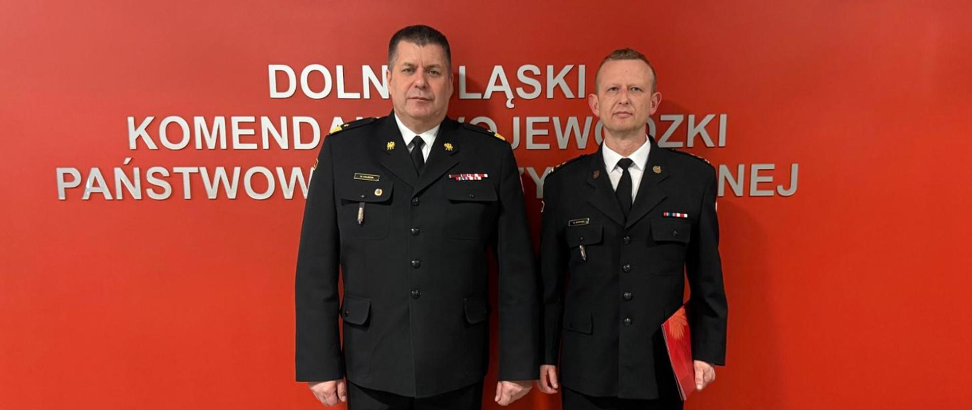 Komendant Wojewódzki i Komendant Powiatowy Państwowej Straży Pożarnej w Lubaniu na tle czerwonego baneru z napisem Dolnośląski Komendant Wojewódzki Państwowej Straży Pożarnej podczas uroczystości wręczenia nominacji na Komendanta Powiatowego Państwowej Straży Pożarnej w Lubaniu. Komendanci są w mundurach wyjściowych.