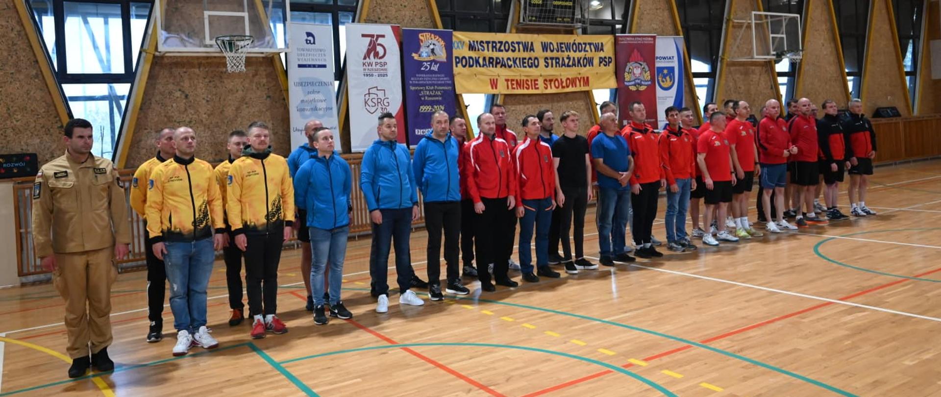Na zdjęciu widać grupę osób stojących w rzędzie w hali sportowej. Uczestnicy ubrani są w różnokolorowe stroje sportowe — żółte, niebieskie, czerwone i czarno-czerwone bluzy oraz dresy. Nad nimi wiszą banery informujące o wydarzeniu:
„XXV Mistrzostwa Województwa Podkarpackiego Strażaków PSP w Tenisie Stołowym”.