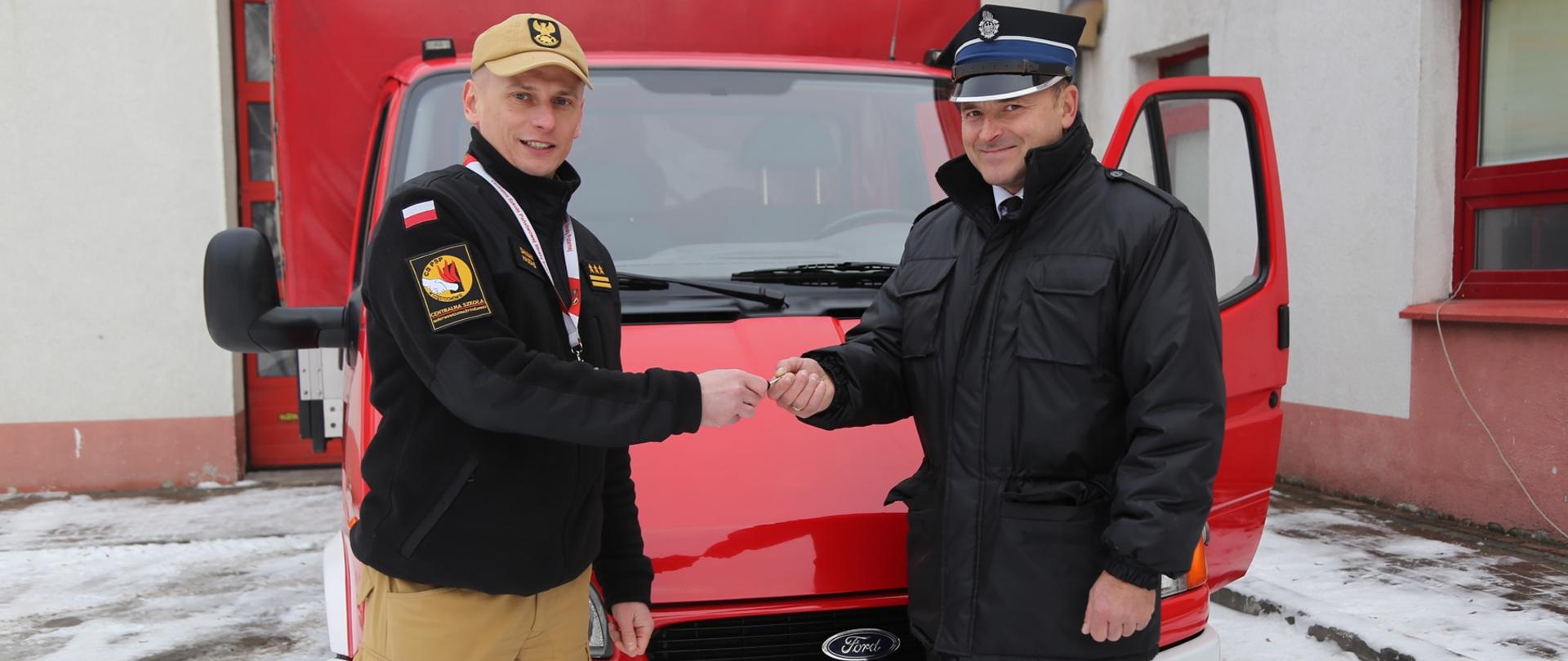 Komendant Centralnej Szkoły PSP przekazuje kluczyki do samochodu druhowi OSP, w tle przekazywany samochód marki Ford Transit