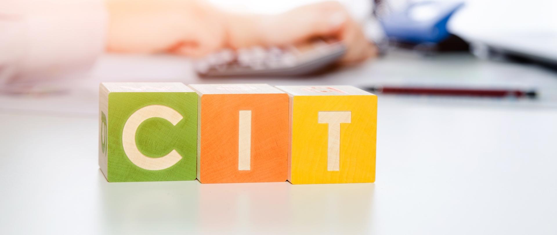 Kolorowe kostki z napisem CIT a w tle rozmyte dłonie i kalkulator