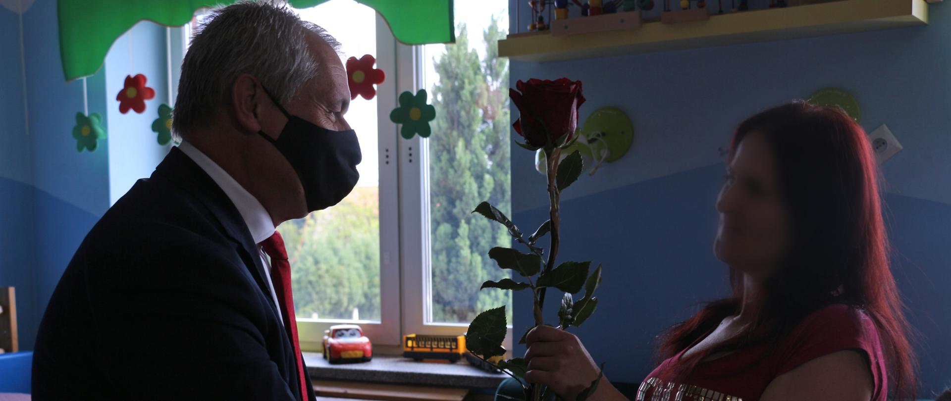 Na zdjęciu wojewoda, stojący po lewej stronie kadru, wręczający czerwoną różę kobiecie, która stoi na przeciw niego. Wojewoda ubrany w ciemny garnitur, zwrócony jest bokiem do kadru. Twarz kobiety została zamazana - z uwagi na ochronę jej wizerunku. Zdjęcie zrobione pod światło. W tle wystrój pokoju, w którym zrobiono zdjęcie. Po lewej stronie widać granatową ścianę, fragment kanapy z różowym pledem. 