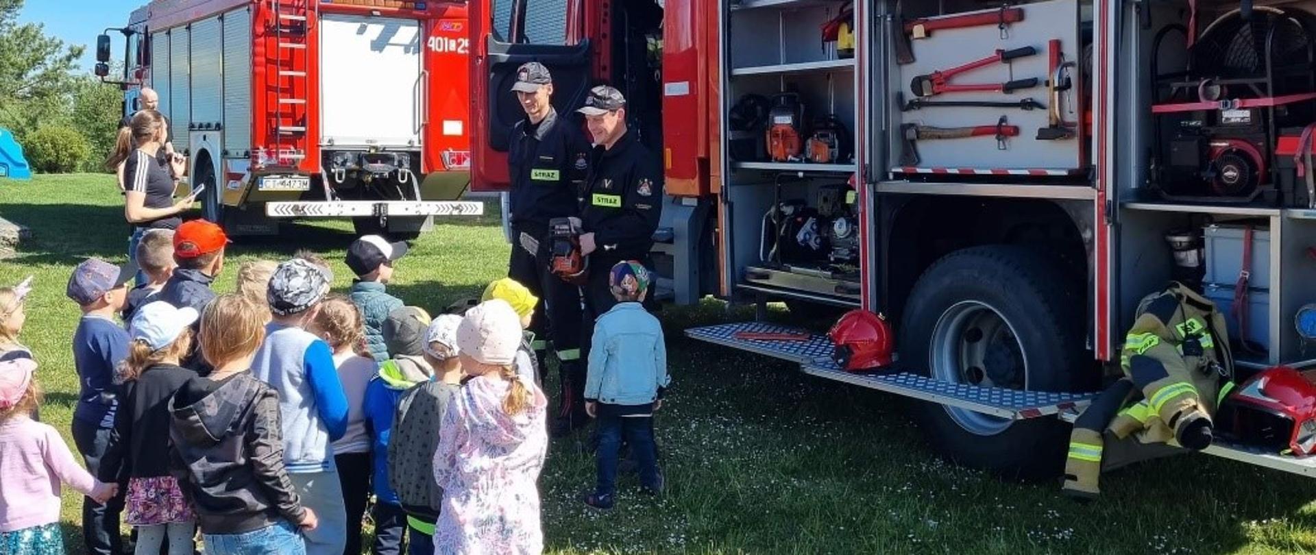 Zdjęcie przedstawia czerwony samochód pożarniczy z uniesionymi roletami, które odsłaniają skrytki ze sprzętem ratowniczo-gaśniczym. Przed pojazdem stoi dwóch funkcjonariuszy straży pożarnej ubranych w czarne umundurowanie koszarowe. Funkcjonariusze pokazują i wyjaśniają działanie sprzętu, jaki znajduje się w skrytkach wozu pożarniczego. Wokół strażaków zgromadzonych jest 15 dzieci oraz ich opiekun. W tle widać tył drugiego pojazdu pożarniczego oraz część budynku szkoły. Prezentacja sprzętu odbywa się na dworze, wszyscy stoją na zielonym trawniku. Pogoda jest słoneczna, niebo jest koloru jasno niebieskiego bez zachmurzenia.