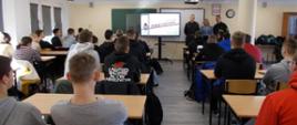 Zdjęcie z tyłu sali szkolnej. Widoczni są uczniowe siedzący w rzędach i przysłuchujący się prowadzącym zajęcia strażakom