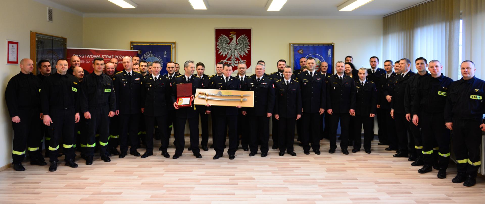 Na zdjęciu widzimy funkcjonariuszy na zbiórce. Zbiórka ma miejsce w KM PSP w Tarnowie. Zbiórka zorganizowana jest z okazji przejścia na zaopatrzenie emerytalne funkcjonariuszy z KM PSP w Tarnowie. Funkcjonariusze ubrani są w umundurowanie galowe i specjalne. Stoją na tle godła państwowego.