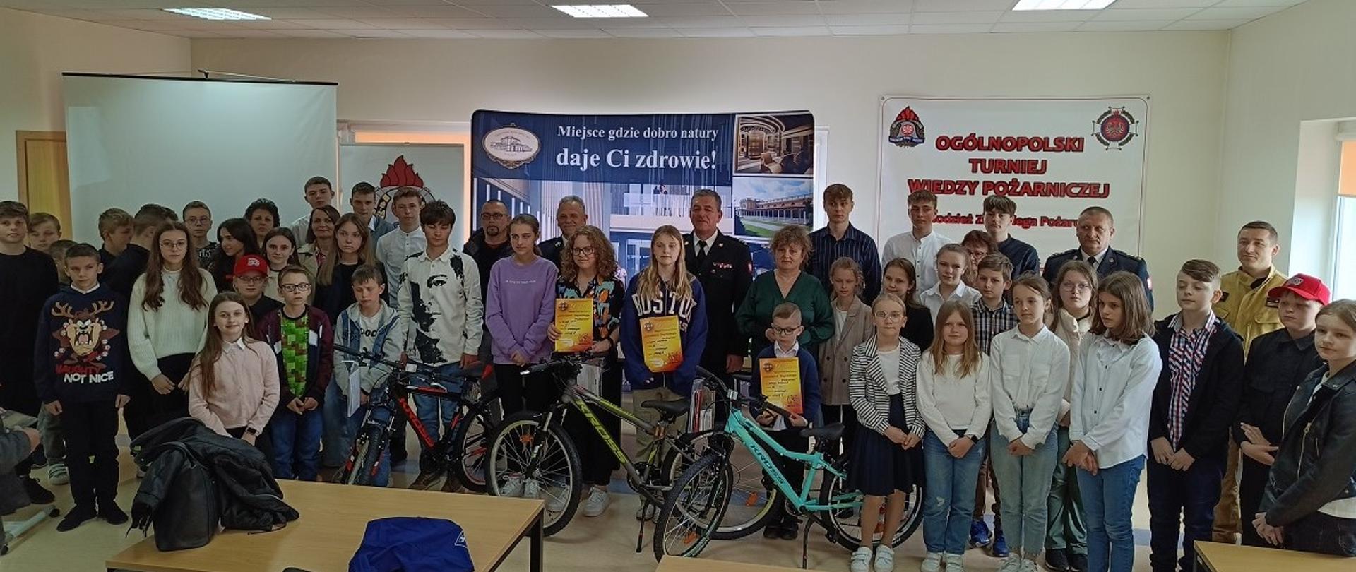 Zdjęcie grupowe uczestników turnieju wraz z nagrodami oraz prowadzącym mł. bryg. Piotrem Dziedzicem. W tle banery . 