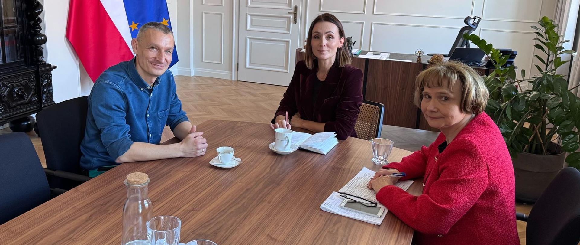 trzy osoby, w tym dwie kobiety, siedza przy stole i rozmawiają