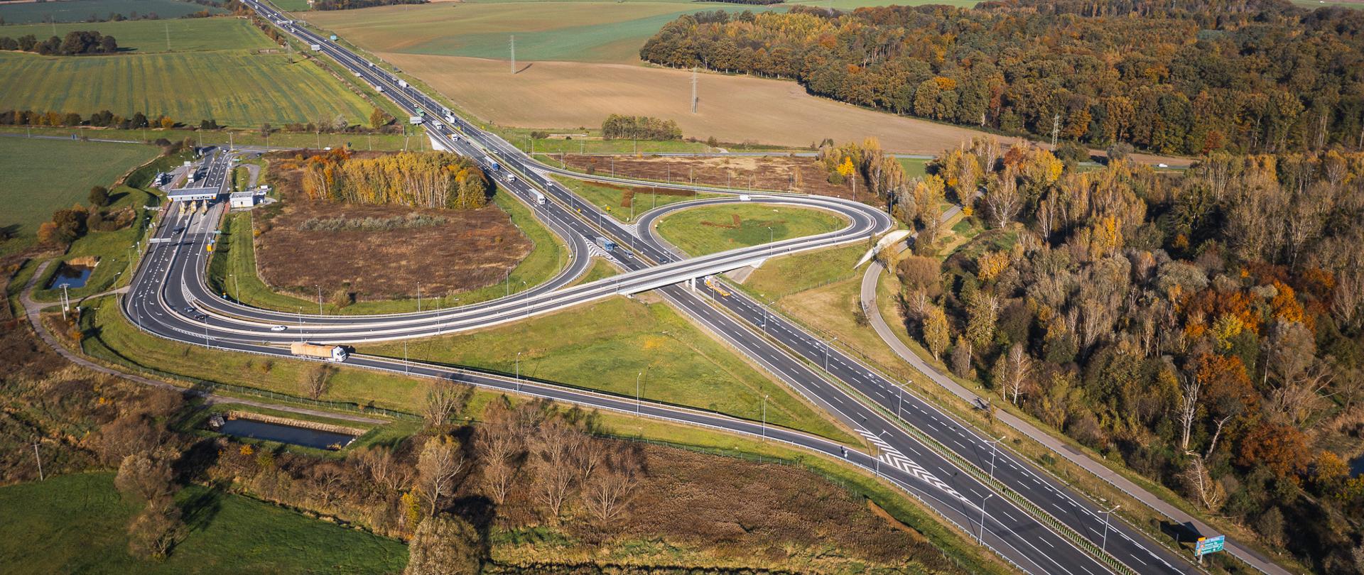 Widok z lotu ptaka na węzeł drogowy z estakadą nad autostradą. Wokół węzła znajdują się pola uprawne, tereny zielone oraz las o jesiennych barwach. Na drogach widoczne są pojazdy poruszające się po autostradzie i łącznicach.
