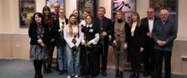 Zdjęcia przedstawia grupę słuchaczy i nauczycieli pozujących na tle kolorowych zdjęć podczas wystawy fotograficznej Światło z Bóbrki. Kobiety trzymają w dłoniach czerwone róże. Z góry pada na nich światło.
