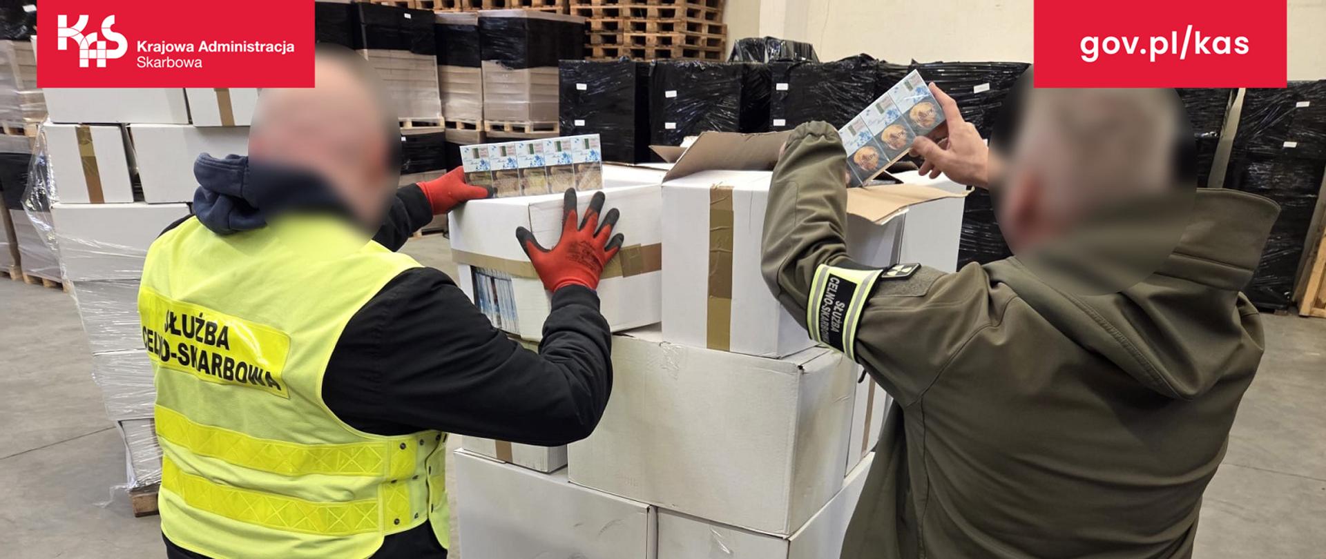 Hala magazynowa, na podłodze stoi paleta z białymi pudłami. Z górnych pudełek dwóch funkcjonariuszy wyjmuje opakowania z papierosami. Na drugim planie wiele palet ustawionych pod ścianami.