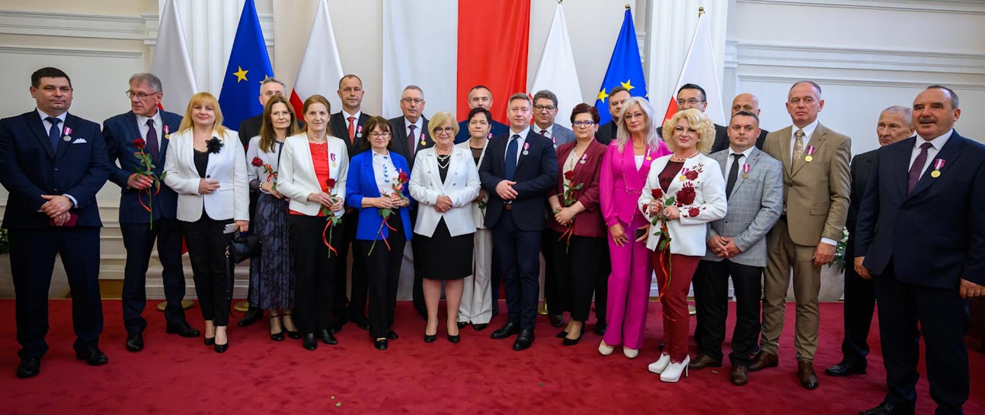 Grupa osób stojąca na czerwonym dywanie na tle flag Polski i UE