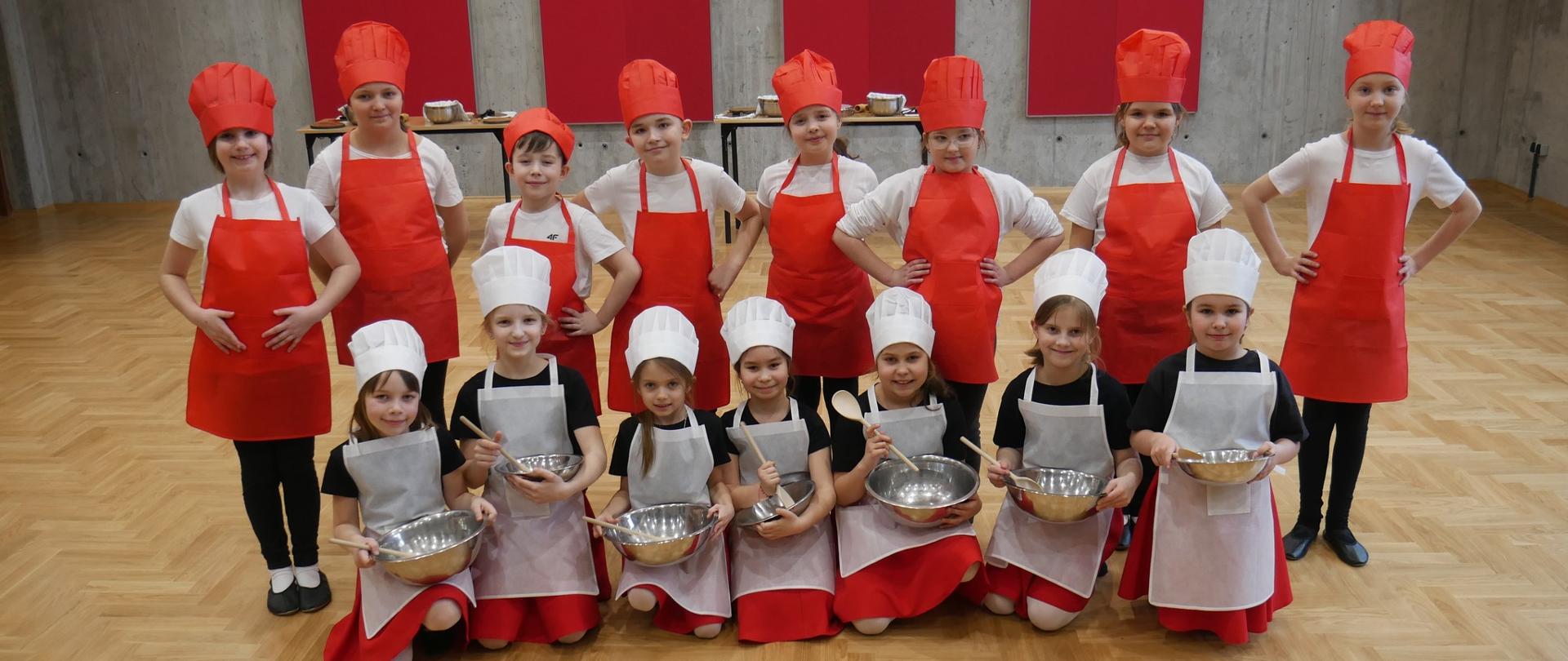 Grupa dzieci w czerwonych kucharskich czapkach i fartuszkach siedzą i stoją na podłodze sali koncertowej szkoły muzycznej, w rękach trzymają miski i łyżki , w tle stoliki z instrumentami muzycznymi