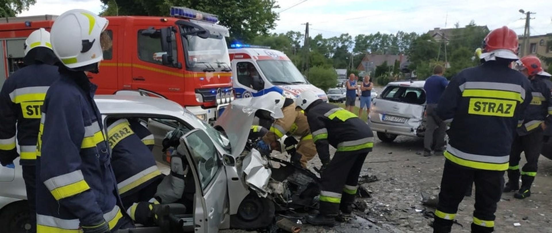 Zdjęcie przedstawia służby ratownicze w trakcie prowadzonych działań ratowniczych. Na zdjęciu samochód po kolizji, strażacy, zniszczony pojazd.