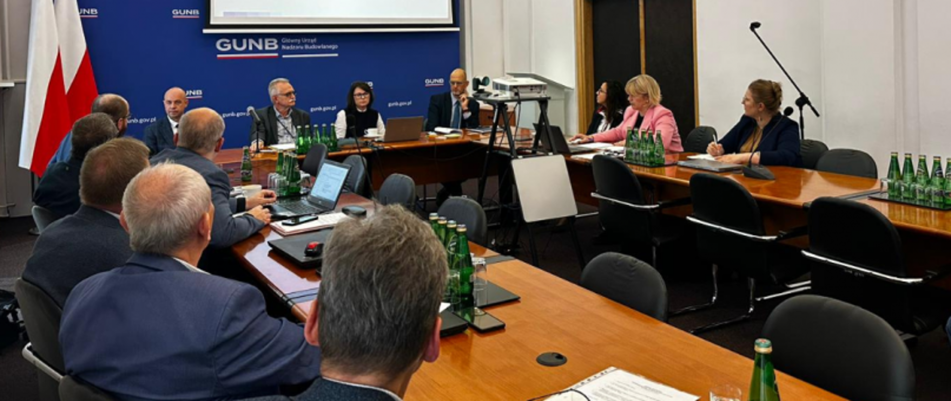 Conference room with people seated around a large wooden table, laptops, documents, and water bottles; blue wall with "GUNB Główny Urząd Nadzoru Budowlanego" text and logos, projector screen, and two flags on the left.