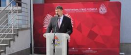Zdjęcie przedstawia Ministra, który stoi przy mównicy na placu przed budynkiem komendy podczas uroczystych obchodów dnia strażaka. 