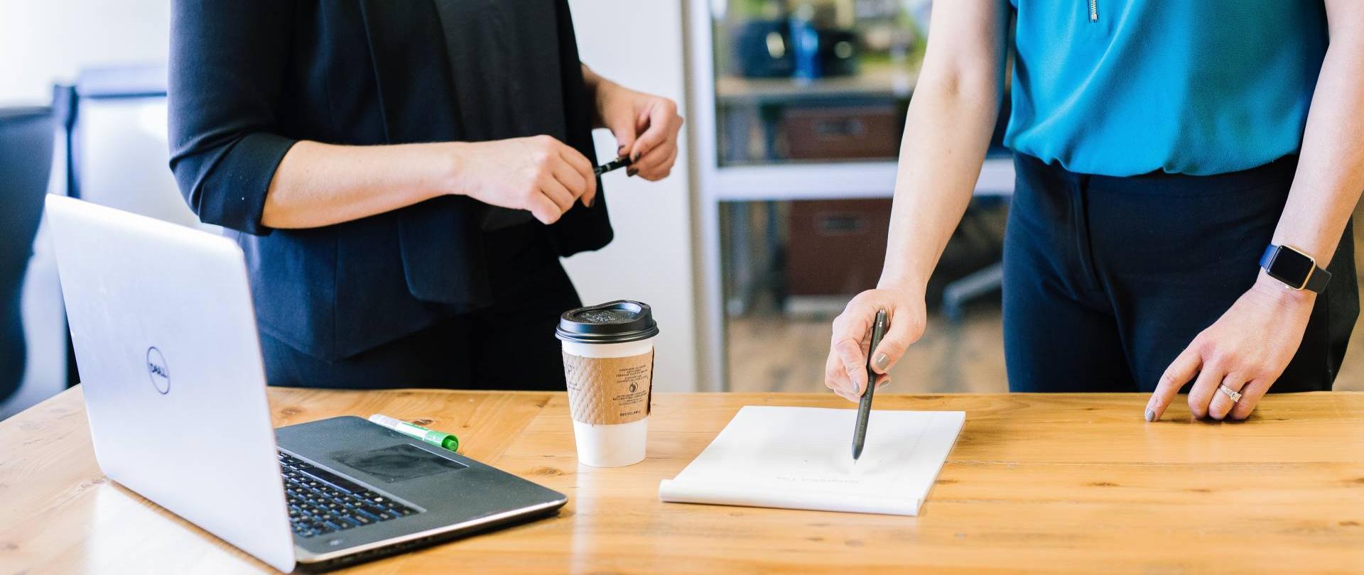fotografia przedstawiająca blat biurka, na którym znajduje się laptop, kubek z kawą i notes, za biurkiem stoją dwie kobiety