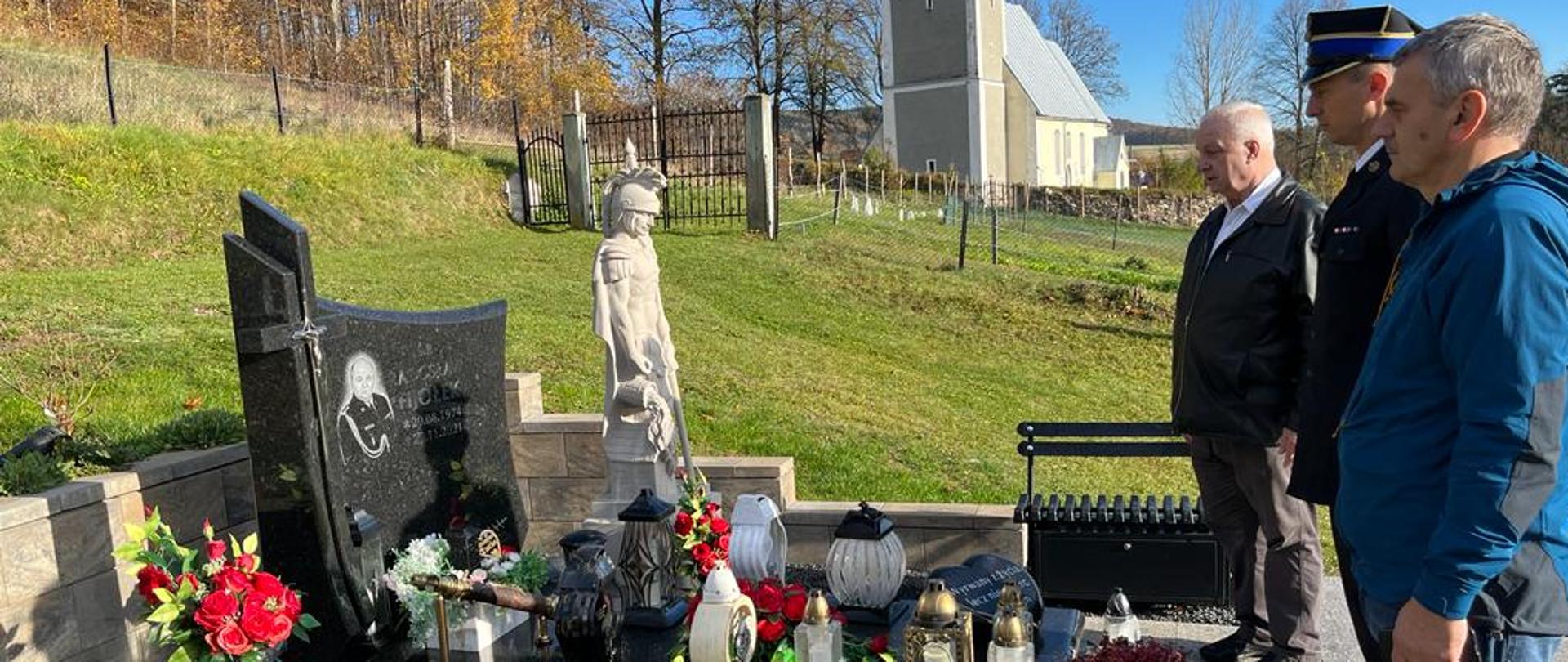 Na zdjęciu widać trzech mężczyzn stojących obok groby zmarłego funkcjonariusza. Zdajecie wykonane na cmentarzu. Grób śp. bryg Radosława Fijołka. 