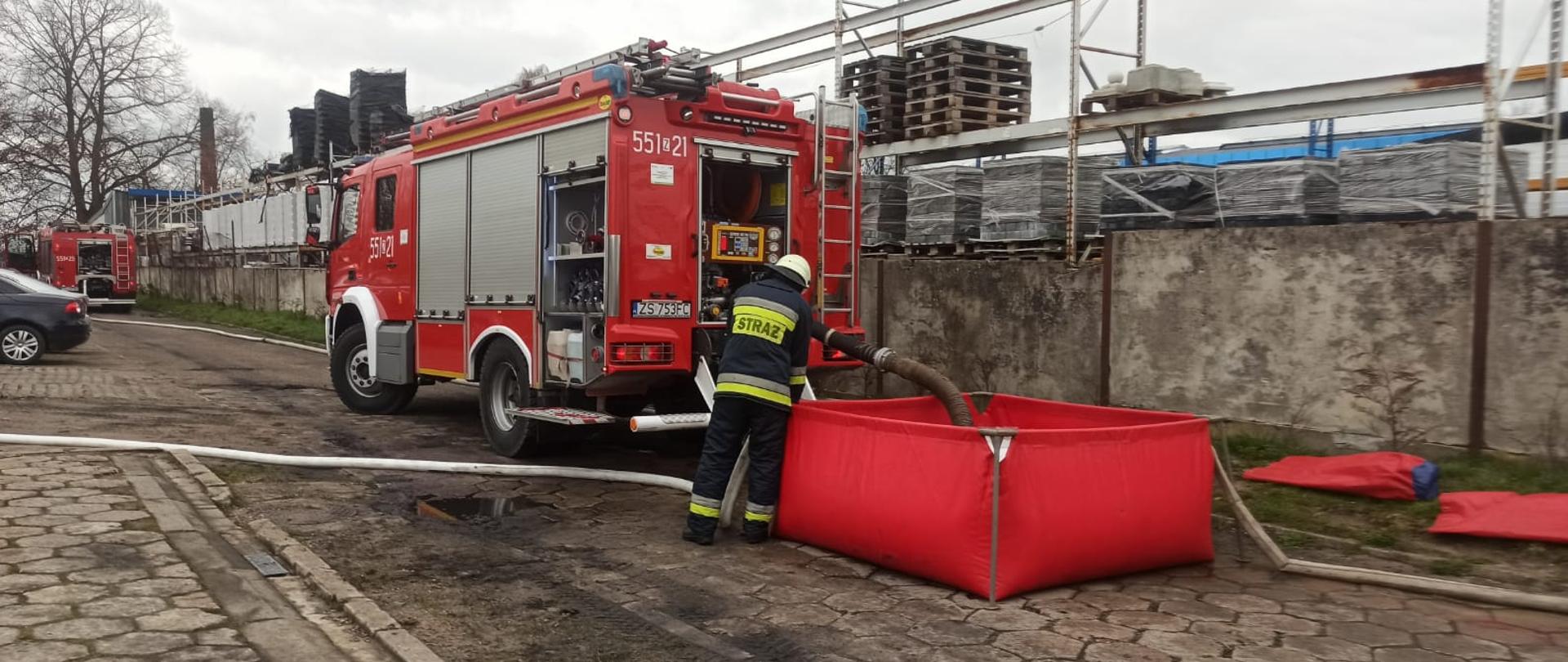 Zdjęcie przedstawia wóz strażacki z podłączonymi wężami, przed nim stoi kolejny wóz strażacki.