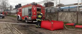 Zdjęcie przedstawia wóz strażacki z podłączonymi wężami, przed nim stoi kolejny wóz strażacki.