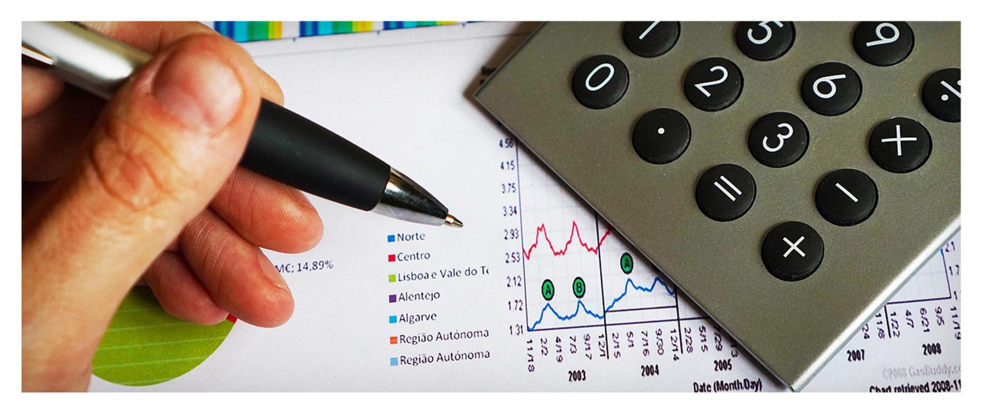 Zdjęcie przedstawia rękę z długopisem oraz wykresy na papierze na którym również znajduje się kalkulator.