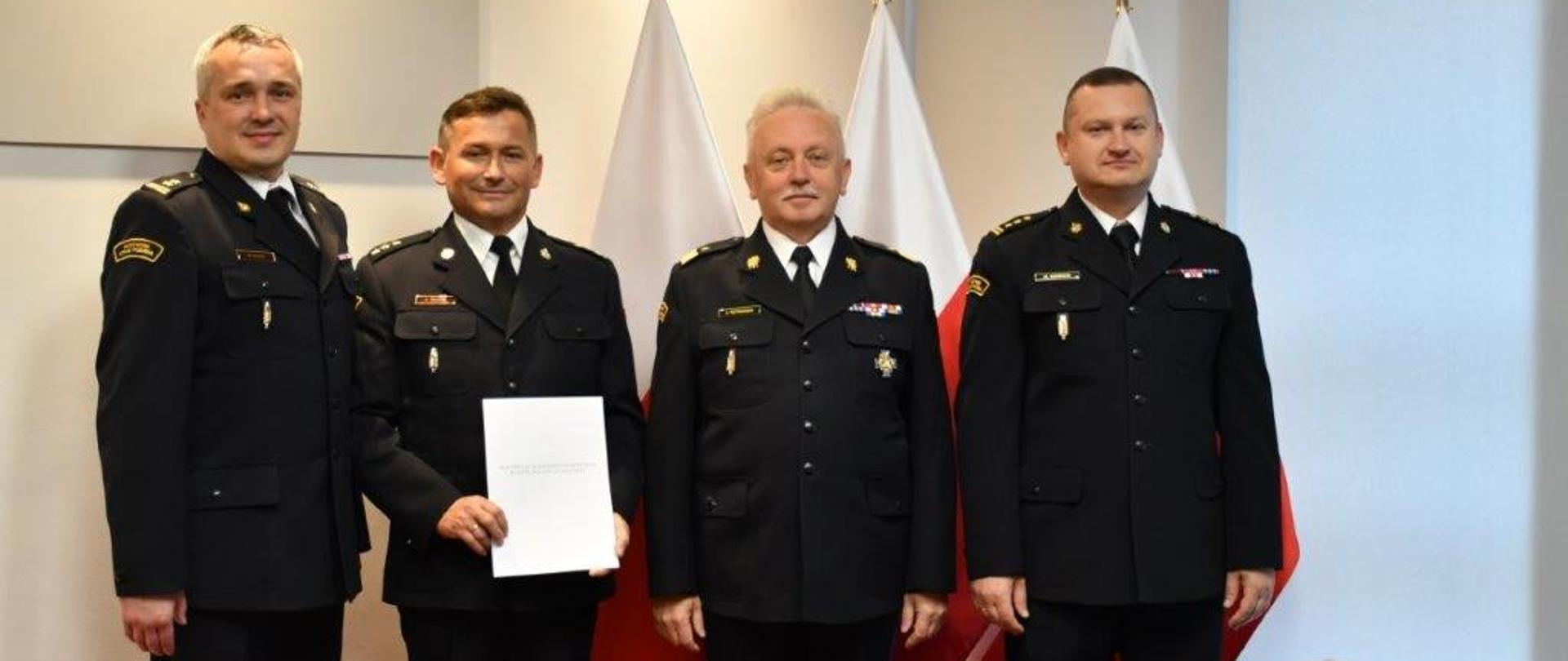 Mazowiecki Komendant Wojewódzki, nadbryg. Jarosław Piotrowski, nowo powołany Zastępca Komendanta Powiatowego PSP w Otwocku, kpt. Grzegorzowi Trzeciakowi, Komendant Powiatowy PSP w Otwocku, bryg. Michał Gigoła oraz Naczelnik Kadr KW PSP st. bryg. Radosław Kozicki stoją w siedzibie Komendy Wojewódzkiej. Za nimi flagi Rzeczypospolitej Polskiej. 