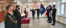 Na zdjęciu w mundurach galowych po lewej stronie Komendant i Zastępca Komendanta Powiatowego a po prawej poczet sztandarowy ze sztandarem. Strażacy składający ślubowanie na środku w ubraniach czarnych koszarowych. Uroczystość ślubowania mam miejsce w sali konferencyjnej.