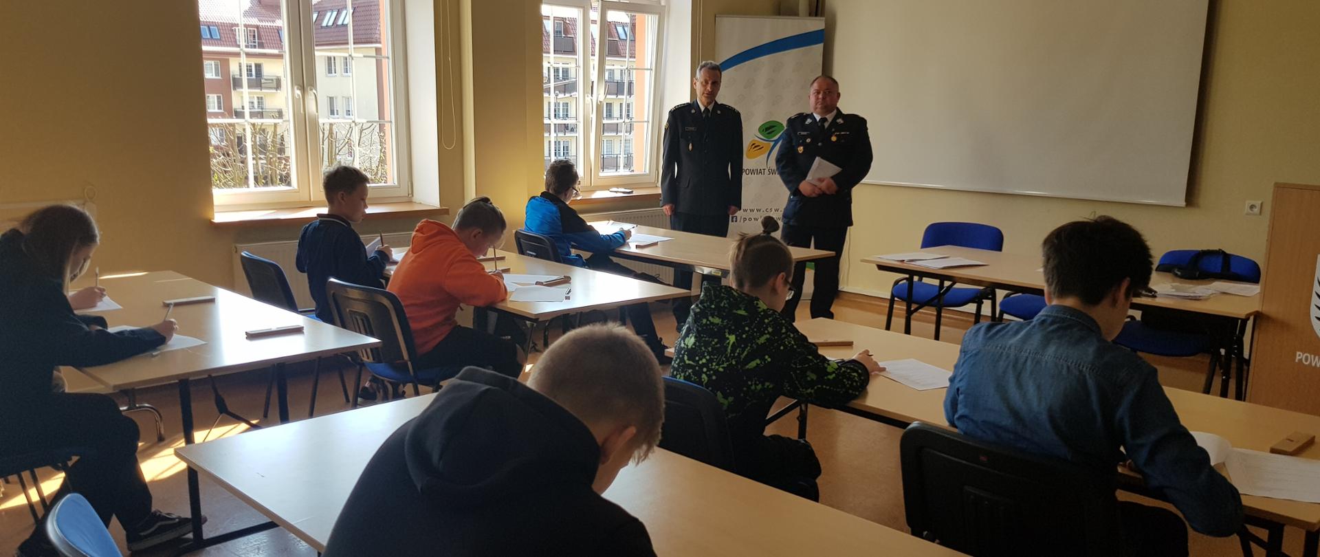 Na zdjęciu widoczni są członkowie komisji konkursowej, Zastępca Komendanta Powiatowego Państwowej Straży Pożarnej w Świeciu st. bryg. Andrzej Rafalski oraz V-ce Przewodniczący Oddziału Powiatowego Związku Ochotniczych Straży Pożarnych dh Maciej Serfin, którzy przyglądają się jak dzieci siedzący w ławkach rozwiązują pytania testowe.