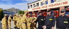 Zastępca Komendanta Głównego PSP wraz z Zastępcą Wielkopolskiego Komendanta Wojewódzkiego witają się z funkcjonariuszami KP PSP Środa Wlkp. W tle budynek komendy i wozy strażackie.