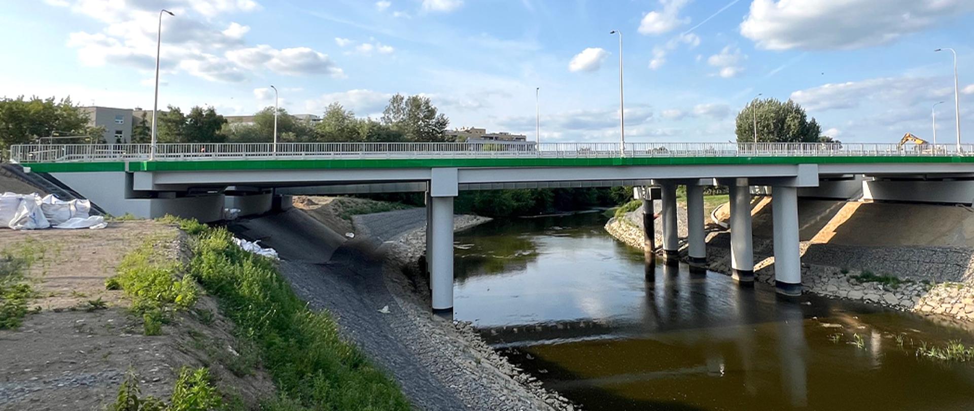 Na zdjęciu widać most wsparty na słupach, budynki mieszkalne i drzewa. Pod mostem płynie niezbyt szeroka rzeka.