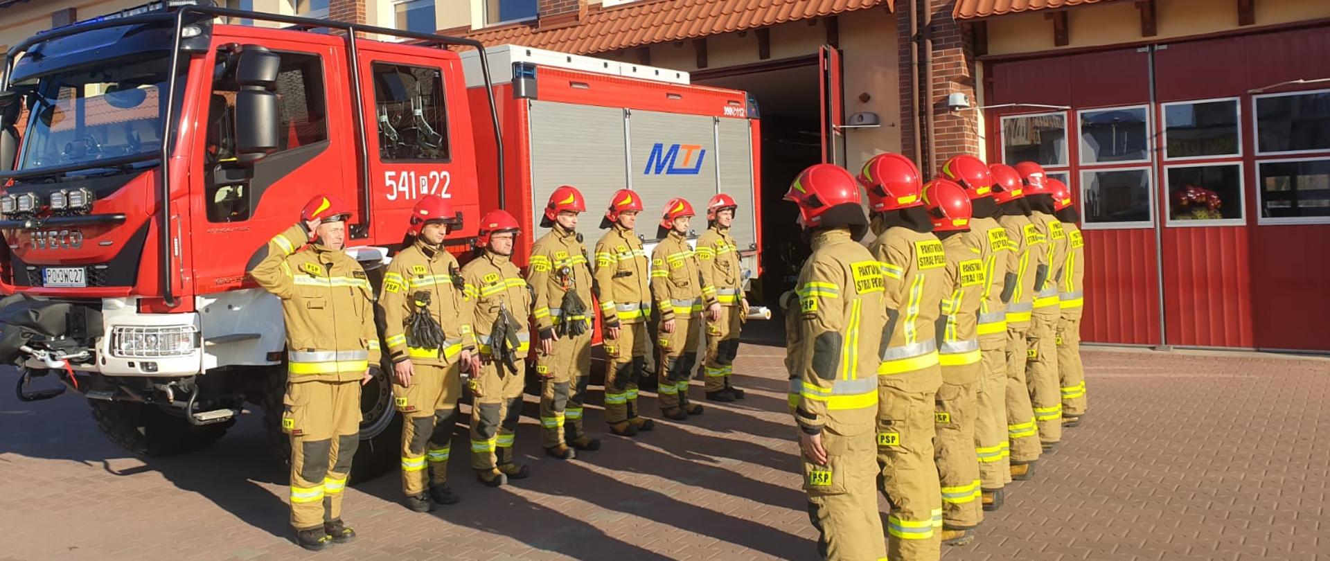 Zdjęcie przedstawia strażaków podczas zmiany służby gdzie uczczono pamięć poległych strażaków na Ukrainie. Na zdjęciu widzimy funkcjonariuszy JRG Ostrzeszów w trakcie zmiany. Zdjęcie wykonane w porze dziennej przed garażami JRG.