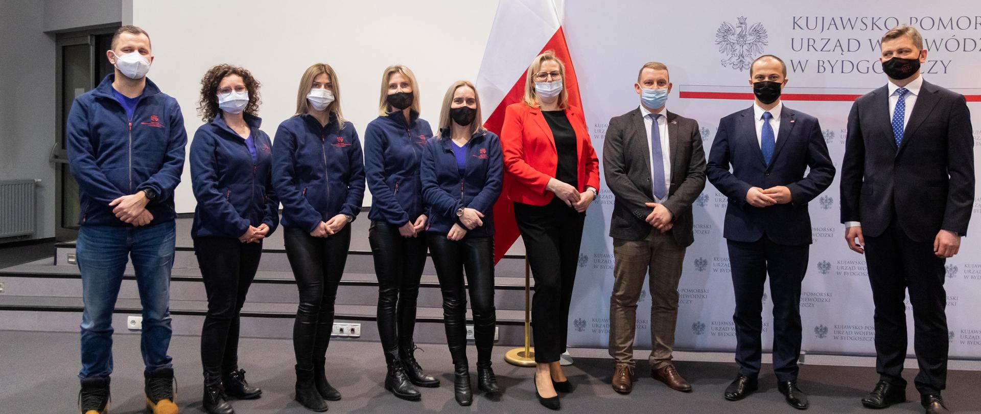9 osób stojących w rzędzie na tle polskiej flagi i barw Kujawsko-Pomorskiego Urzędu Wojewódzkiego