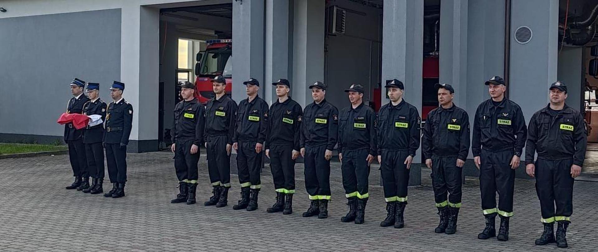 Na zdjęciu widać trzech strażaków, którzy trzymają na rękach flagę Polski. Po prawej stronie stoją strażacy w jednym rzędzie. W sumie trzynaście osób. Wszyscy ubrani są w czarne stroje z jasnożółtymi odblaskami z napisem straż. Na głowach strażacy mają założone czapki z daszkiem. Uroczysta zbiórka odbywa się na placu manewrowym przed budynkiem Komendy Powiatowej Państwowej Straży Pożarnej w Łęczycy. W tle widać budynek komendy. Jest widno. 