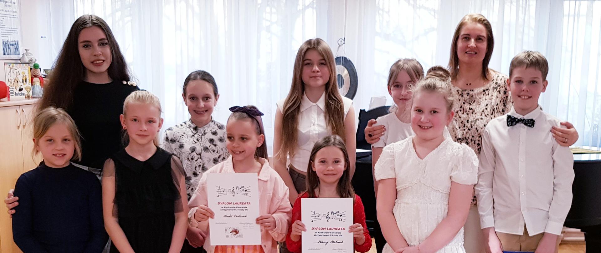 Zdjęcie grupowe wszystkich uczestników konkursu trzymającymi dyplomy wraz z nauczycielem klasy skrzypiec na tle sali rytmiki Państwowej Szkoły Muzycznej I st. w Turku