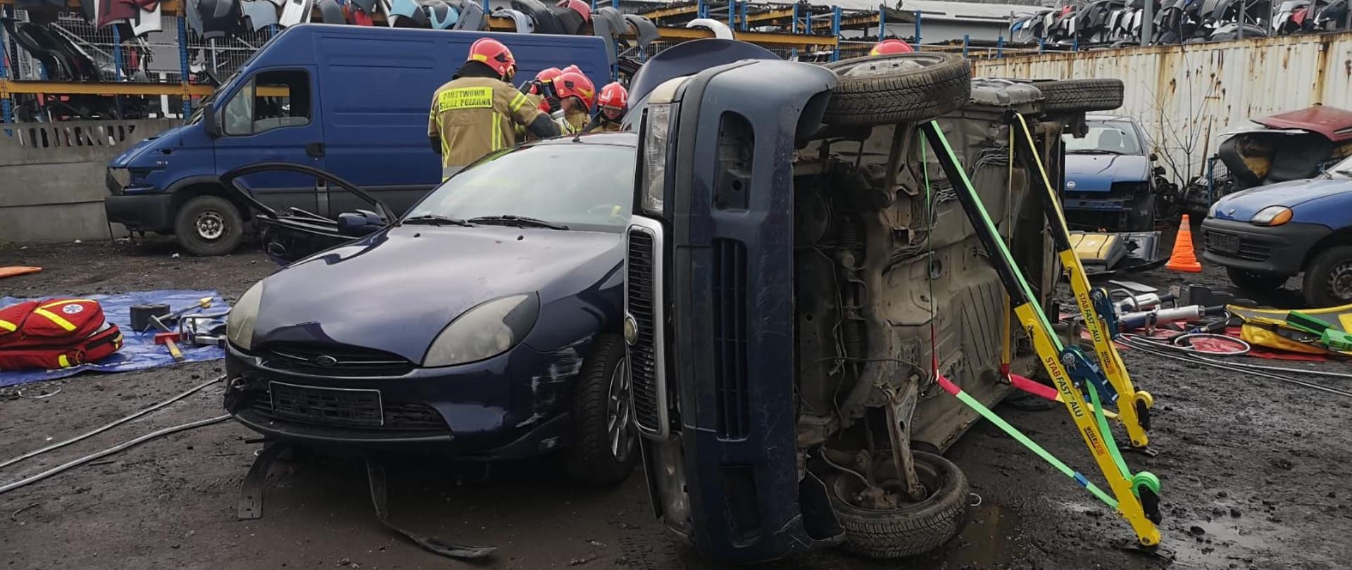 Zdjęcie przedstawia leżący na boku samochód osobowy ustabilizowany podporami, który dachem opiera się o stojący obok samochód.