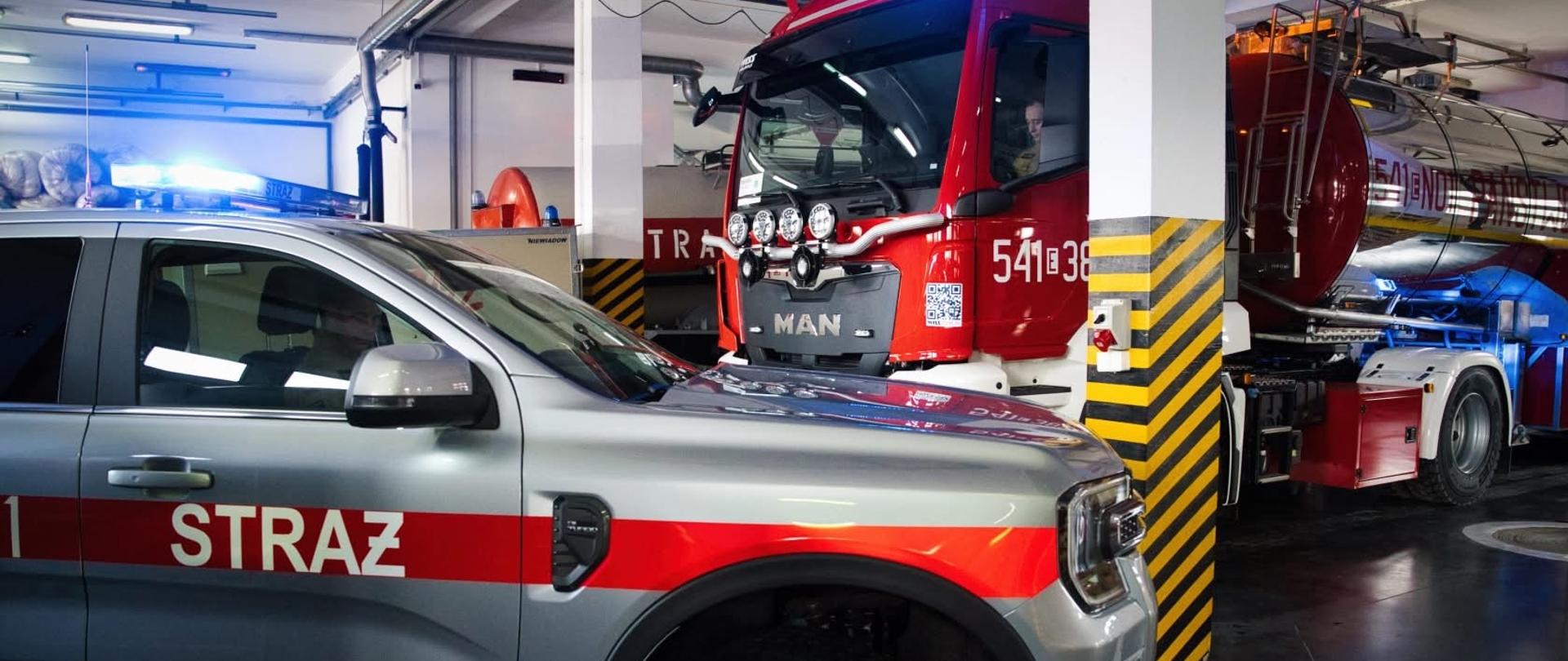 Wnętrze garażu jednostki straży pożarnej, w którym zaparkowany jest nowoczesny srebrny samochód operacyjny marki Ford z napisem „STRAŻ”. Tuż za nim stoi duży, czerwony wóz gaśniczy marki MAN oraz ciężka cysterna, oba z włączonymi niebieskimi sygnałami świetlnymi.