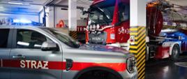 Wnętrze garażu jednostki straży pożarnej, w którym zaparkowany jest nowoczesny srebrny samochód operacyjny marki Ford z napisem „STRAŻ”. Tuż za nim stoi duży, czerwony wóz gaśniczy marki MAN oraz ciężka cysterna, oba z włączonymi niebieskimi sygnałami świetlnymi.