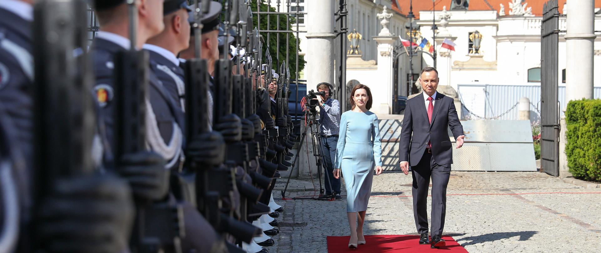 Na zdjęciu znajduje się szpaler, który tworzą umundurowani funkcjonariusze Policji. Wzdłuż szpaleru zmierzają w kierunku fotografa elegancko ubrani kobieta i mężczyzna. 