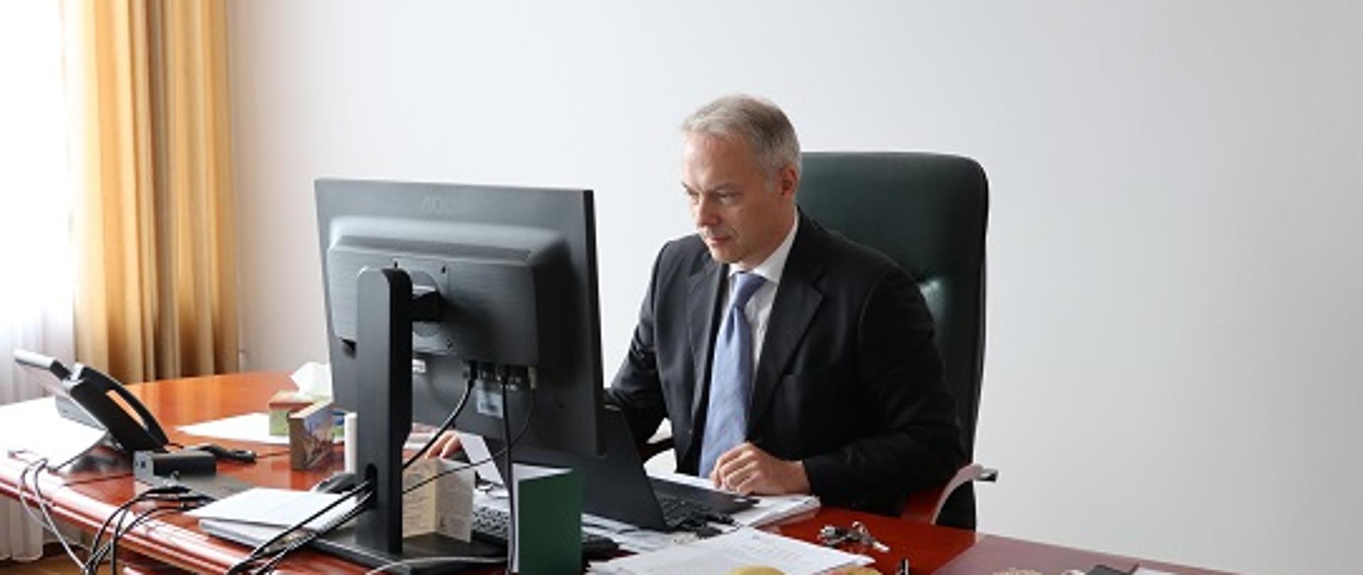 Wiceminister Jacek Żalek siedzi przy biurku, przed komputerem. Na biurku widoczne dokumenty.