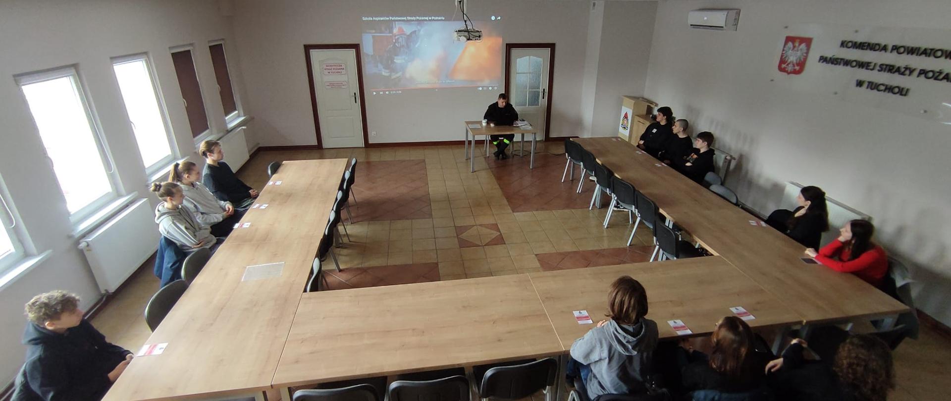 Przy stołach dookoła siedzą uczniowie. Naprzeciwko uczniów siedzi funkcjonariusz PSP. Za funkcjonariuszem PSP widnieje obraz wyświetlony przez rzutnik. Na obrazie widać strażaka w czerwonym hełmie. W tle po lewej i prawej stronie są drzwi. Po lewej stronie widać okna. Po prawej stronie widać godło Polski oraz napis: Komenda Powiatowa Państwowej Straży Pożarnej w Tucholi. Po prawej stronie w rogu widać mównicę, a na ścianie biały klimatyzator. Ściany są koloru białego. Na podłodze są płytki koloru żółtego i pomarańczowego. 