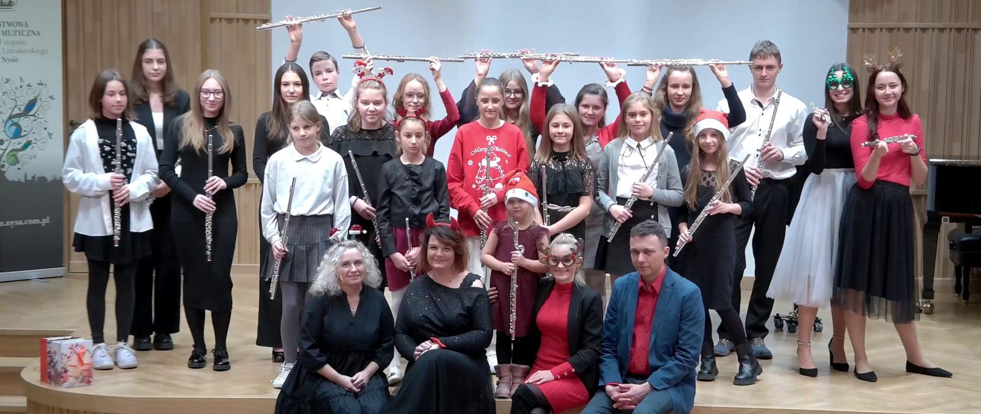 Zdjęcie przedstawia wszystkich wykonawców Mikołajkowego Koncertu Fletowego, który odbył się w Sali Koncertowej im. Jerzego Kozarzewskiego w PSM I i II stopnia im. Witolda Lutosławskiego w Nysie 9 grudnia 2022. W tylnym rzędzie uczniowie stoją z uniesionymi do góry fletami. W środkowym rzędzie uczniowie trzymają flety w rękach. Z przodu siedzą nauczyciele.