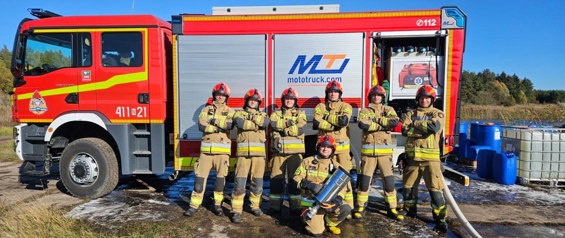 Siedmiu strażaków w mundurach bojowych przy dużym czerwonym samochodzie strażackim. Słonecznie