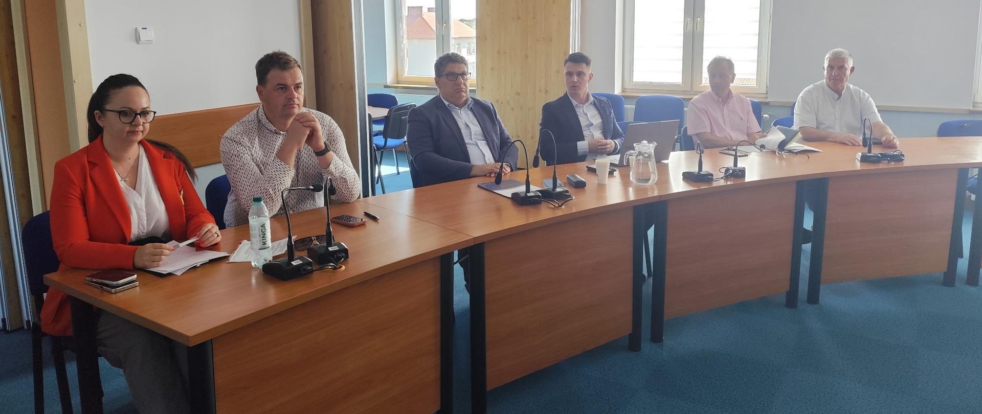 Grupa osób siedzi w jasnym pomieszczeniu przy dużym drewnianym biurku. Przed nimi stoją mikrofony. W tle są okna.