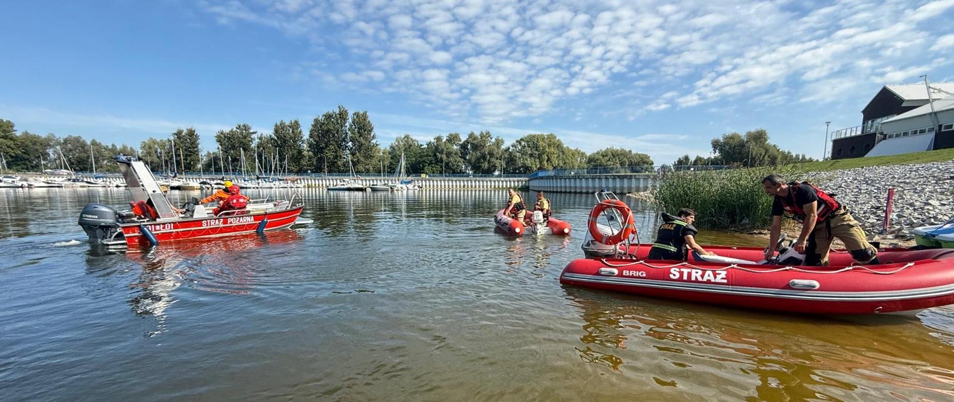 trzy łodzie strażackie ze strażakami w środku są na wodzie, w tle widać inne łódki które stoją w porcie