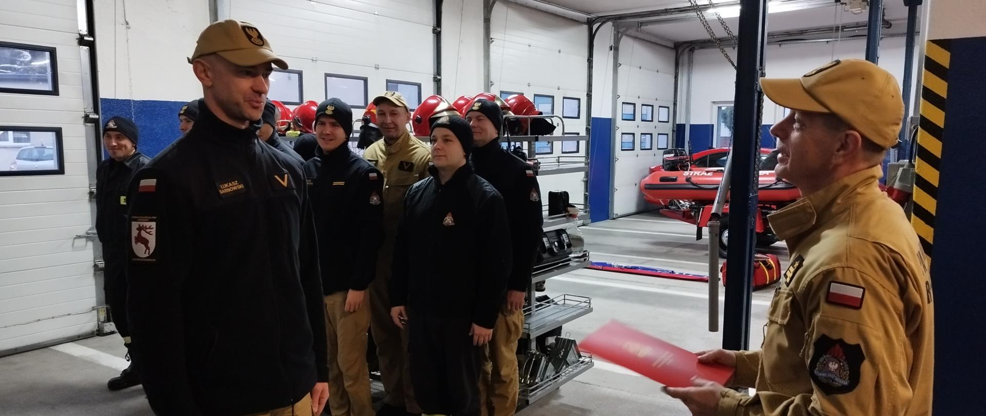 Zdjęcie przedstawia st. bryg. Andrzeja Góreckiego Komendanta Powiatowego PSP w Brodnicy wręczającego ogn. Łukaszowi Sarnowskiemu list gratulacyjny z okazji jubileuszu 20 lat pracy i służby. W tle strażacy podziału bojowego JRG Brodnica.