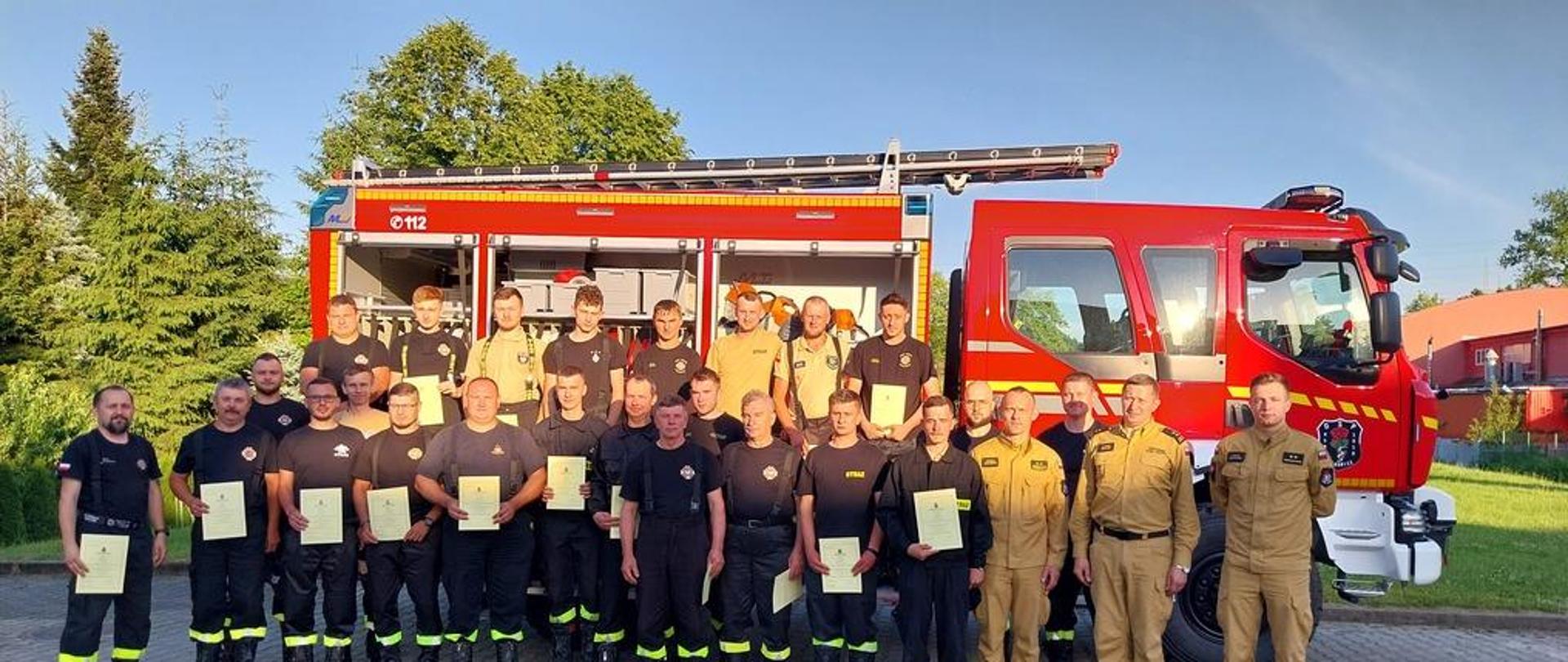 Na zdjęciu na tle samochodu pożarniczego na placu Komendy Powiatowej Państwowej Straży Pożarnej w Gorlicach strażacy z Ochotniczych Straży Pożarnych i członkowie Komisji egzaminacyjnej strażacy z Państwowej Straży Pożarnej po egzaminie kończącym szkolenie kierowców – konserwatorów sprzętu ratowniczego OSP. Strażacy ubrani w ubrania koszarowe koloru czarnego i piaskowego bez czapek.