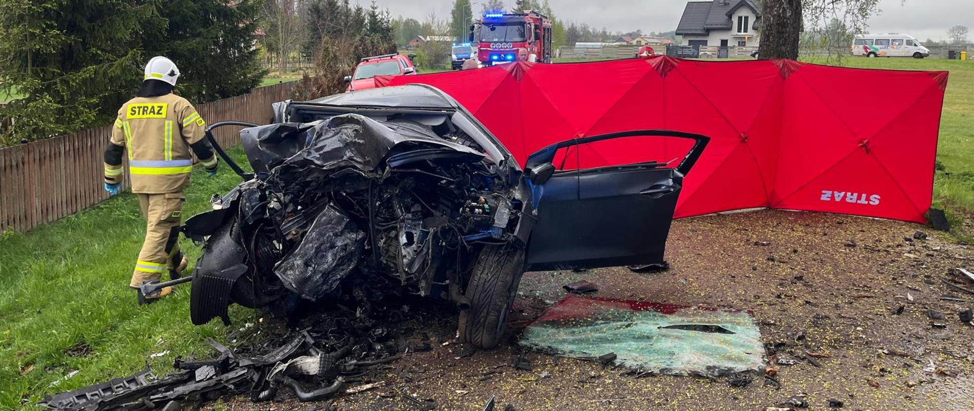Zdjęcie przedstawia wrak pojazdu osobowego po zderzeniu z drzewem, a po jego lewej stronie widać strażaka Ochotniczej Straży Pożarnej w Grabowie. Następnie można zaobserwować parawan ochronny do zabezpieczenia miejsca zdarzenia koloru czerwonego. W oddali widać również pojazd pożarniczy oraz radiowóz z włączony sygnałami świetlnymi koloru niebieskiego. 
