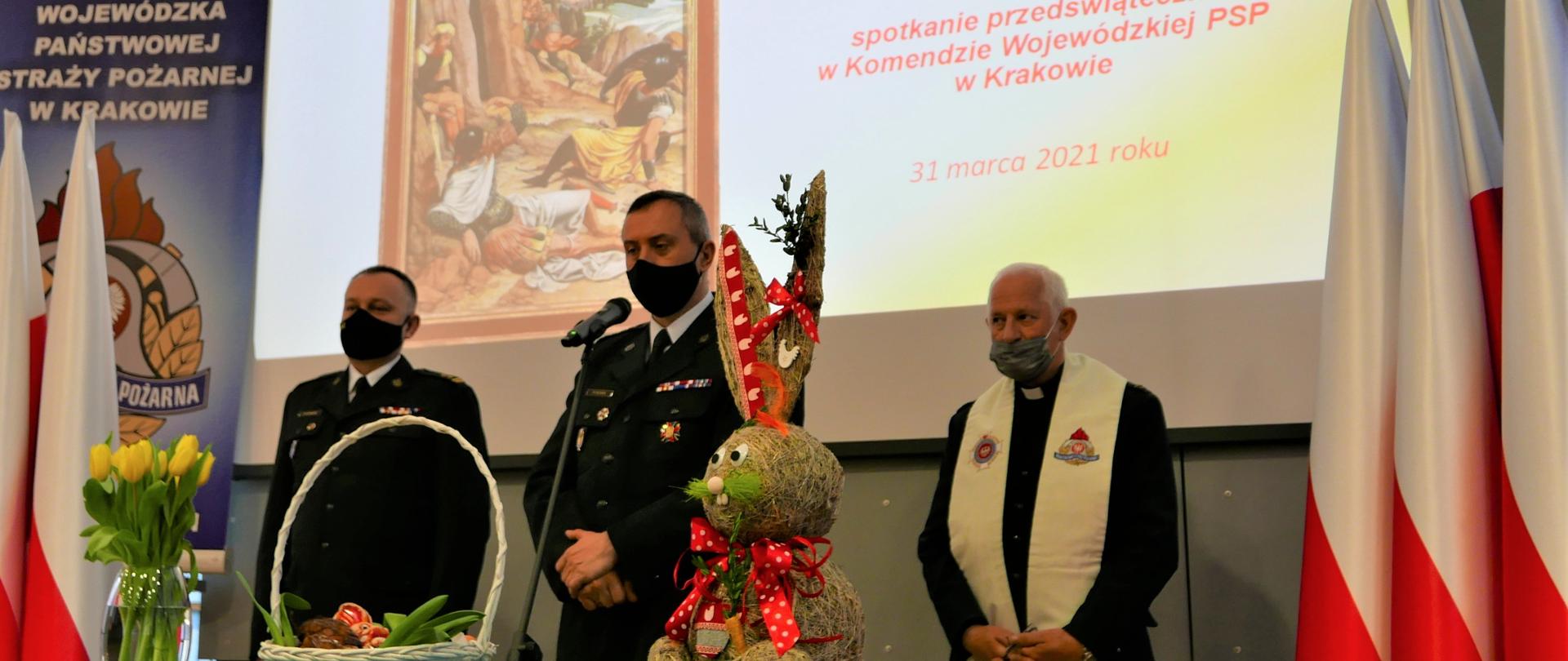 Komendant Wojewódzki PSP wraz z Zastępcom i Kapelanem stoją na tle slajdu z motywem wielkanocnym. Przed nimi na stole wazon z żółtymi tulipanami, króliczek i koszyczek wielkanocny. Po bokach flagi Polski.