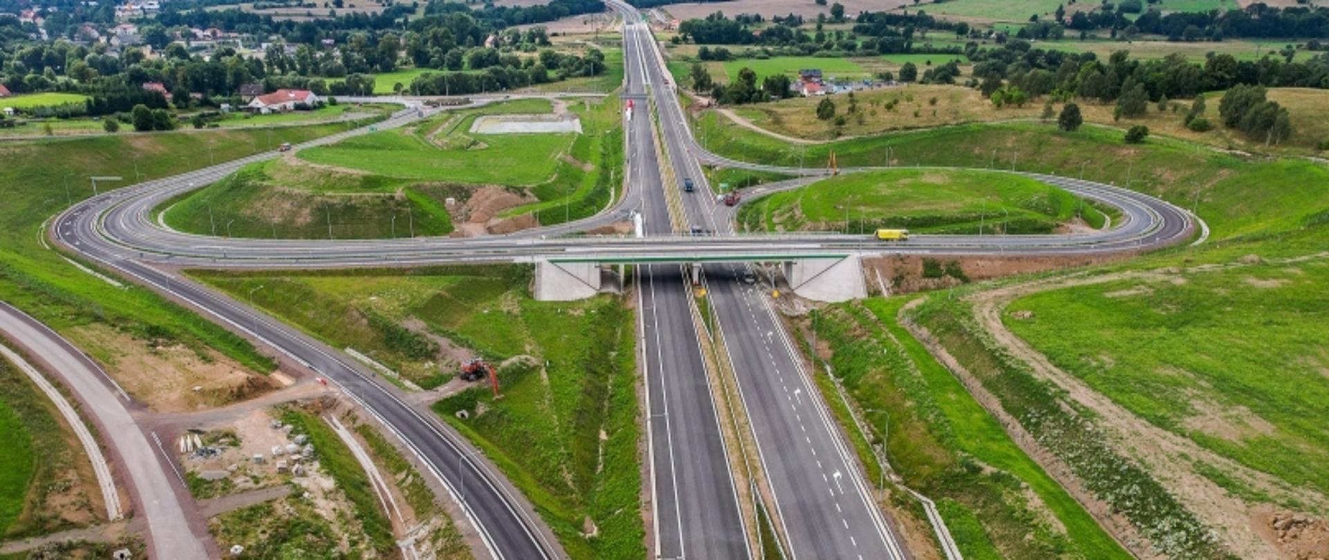 Węzeł drogowy z estakadami i zjazdami w otoczeniu pól i zabudowy wiejskiej, widziany z lotu ptaka.