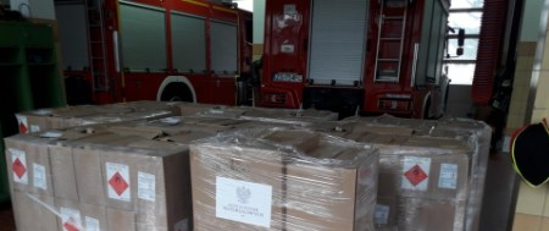 Na zdjęciu stoją kartony ze środkami ochrony indywidualnej na paletach a w tle widać wozy strażackie.