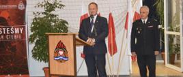 Wręczenie promes. Sala sportowa w Szkaradowie. Przy mównicy przemawia do zebranych Pan starosta Adam Sperzyński. Z przodu na mównicy umieszczony jest logotyp PSP. Obok przemawiającej stoi strażak PSP w umundurowaniu wyjściowym. W tle flagi państwowe, kwiat oraz materiały promujące KP PSP w Rawiczu i OSP w Szkaradowie.