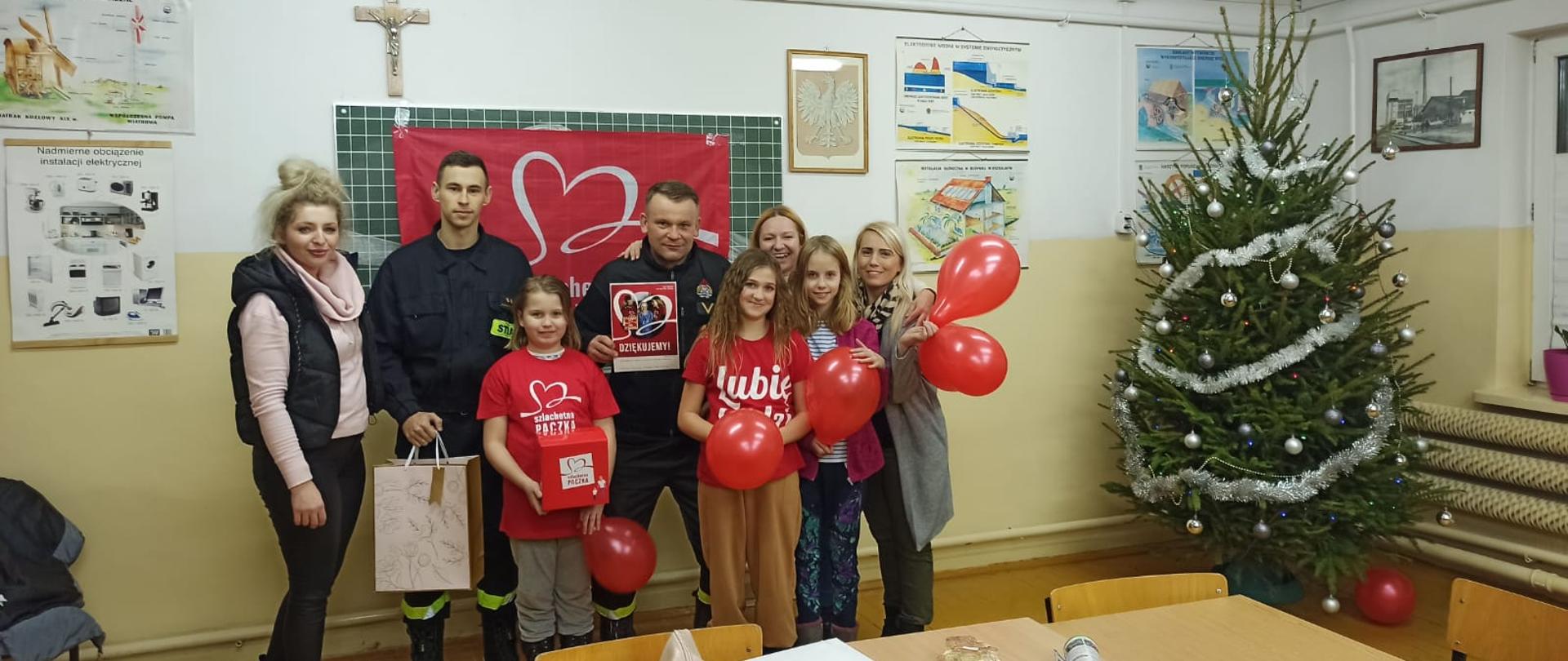 W sali lekcyjne stoi kilka osób z czerwonymi balonikami. Wśród nich dwóch strażaków w mundurach. Obok choinka.